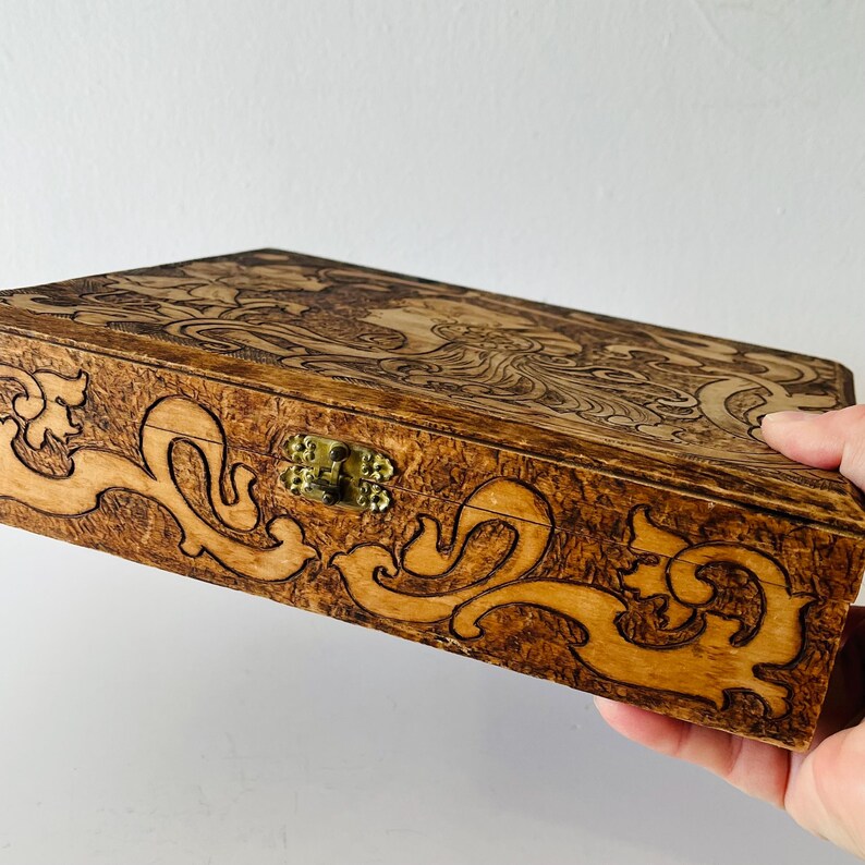 May include: A rectangular wooden box with intricate carved designs. The box features a brass clasp and is held by a hand. The carvings include floral and swirling patterns, creating a decorative and detailed appearance. The wood has a warm, natural tone.