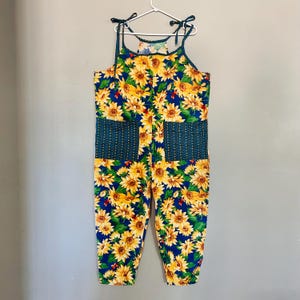 Peut inclure: Combinaison une pièce avec un imprimé tournesol vibrant sur fond bleu. La combinaison comporte des bretelles réglables, deux poches avant avec un motif à pois turquoise et une coupe décontractée. Le vêtement est suspendu à un cintre blanc.