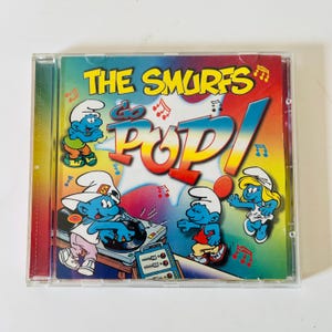 May include: A CD case featuring the title "THE SMURFS Go POP!" in yellow and red text. The cover art includes cartoon Smurfs, musical notes, and a DJ setup. The background is a mix of blue, green, and yellow.