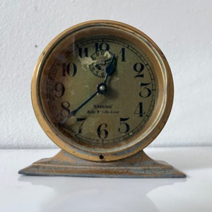 Pode incluir: Um relógio antigo Westclox Baby Ben De Luxe em tom dourado. O relógio tem um mostrador redondo com algarismos e ponteiros pretos e um ponteiro de segundos azul. O relógio assenta numa base retangular.