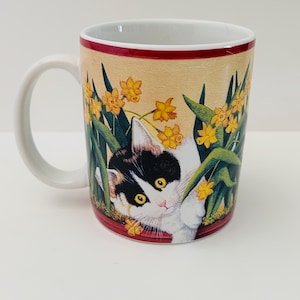 May include: White ceramic mug with a handle, featuring a cat peeking through yellow flowers and green leaves. The mug has a red rim and a beige background. The cat is black and white with yellow eyes.