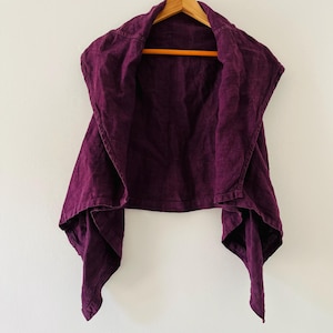 May include: A sleeveless, deep purple linen vest with a draped collar and an asymmetrical hem. The vest is displayed on a wooden hanger against a white wall. The fabric has a textured appearance.