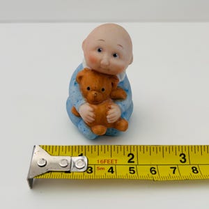 Pode incluir: Uma pequena estatueta de um bebê com uma roupa azul, segurando um ursinho de pelúcia marrom. O bebê tem cabeça careca, bochechas rosadas e olhos azuis. A estatueta tem aproximadamente 5 cm de altura.