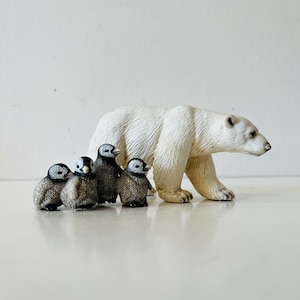 May include: A white polar bear figurine walks alongside four small penguin figurines. The penguins are grey, black, and white. The polar bear has a textured surface and is walking to the right.