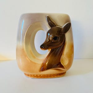Może przedstawiać: Ceramiczny wazon z motywem głowy jelenia. Wazon ma prostokątny kształt z dużym, okrągłym wycięciem. Głowa jelenia jest brązowa i wystaje z boku. Wazon ma gradient kolor&oacute;w, od jasnobrązowego do brązowego.