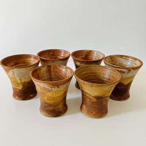 May include: A set of six ceramic tumblers featuring a flared rim and a tapered base. Each vessel displays a variegated glaze in earthy tones of brown, tan, and cream, with visible speckling and a smooth, glossy finish across the exterior surface.