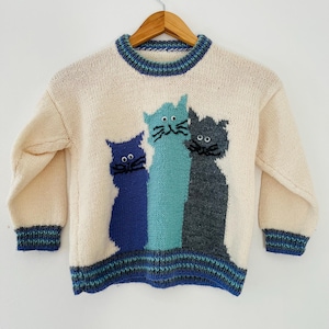 May include: Cream-colored knit sweater featuring three cat silhouettes in blue, teal, and gray. The sweater has a ribbed neckline, cuffs, and hem with blue and teal stripes. The cats have black whiskers and eyes. The sweater is hanging on a wooden hanger.