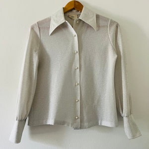 May include: A silver sparkly long-sleeved button-up shirt with a wide collar. The shirt has pearl buttons.