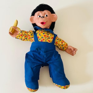 May include: A monkey doll with black hair, a peach-coloured face, and a wide open mouth. The doll wears blue overalls over a yellow long-sleeved shirt with a red and blue floral pattern. It holds a small yellow banana in its right hand.