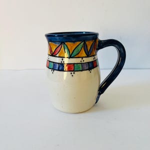 May include: A white ceramic mug with a blue handle and colourful geometric designs around the rim. The design features yellow, red, blue, green, and purple accents.