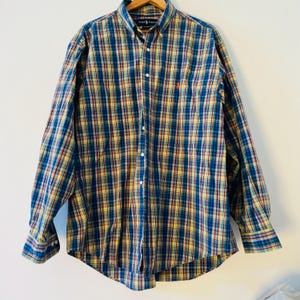 May include: A long-sleeved button-down shirt with a plaid pattern in blue, yellow, and red. The shirt has a button-down collar and a small orange logo on the chest. It is displayed on a wooden hanger.