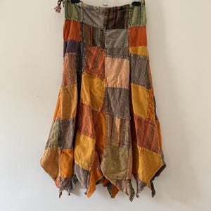 Op de afbeelding: Een patchwork rok in tinten oranje, geel, bruin en groen. De rok heeft een verscheidenheid aan vierkanten stof die aan elkaar zijn genaaid, waardoor een uniek en gestructureerd ontwerp ontstaat. De rok heeft een vloeiende, asymmetrische zoom.