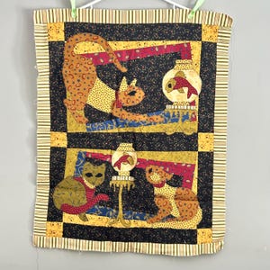 May include: A quilted wall hanging featuring a black background with colorful cats and fish. The cats are in various poses, including sitting, standing, and jumping. The fish are in a bowl with a gold rim.