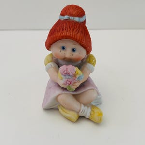 May include: A ceramic figurine of a seated girl with red hair and blue eyes, holding a bouquet of flowers. She wears a pink dress, yellow shoes, and a blue ribbon in her hair. The figurine has a vintage, collectible aesthetic.