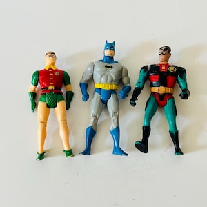 May include: Three action figures of Batman and Robin. Batman is in the centre, wearing a grey and blue suit with a yellow belt and bat symbol. The Robin figures are on either side, in red, green, and yellow costumes.