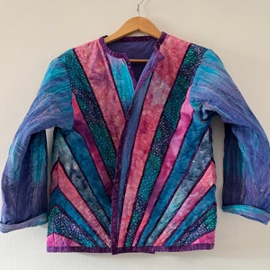 Può includere: Una giacca trapuntata con un design unico che presenta una miscela di tonalità rosa, blu e viola. La giacca ha un collo a V e maniche lunghe.