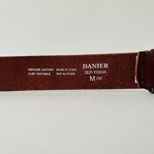 May include: Brown leather belt with white text that reads "GENUINE LEATHER CUIR VERITABLE MADE IN CHINA FAIT AU CHINE DANIER DLP-11300A M/M"