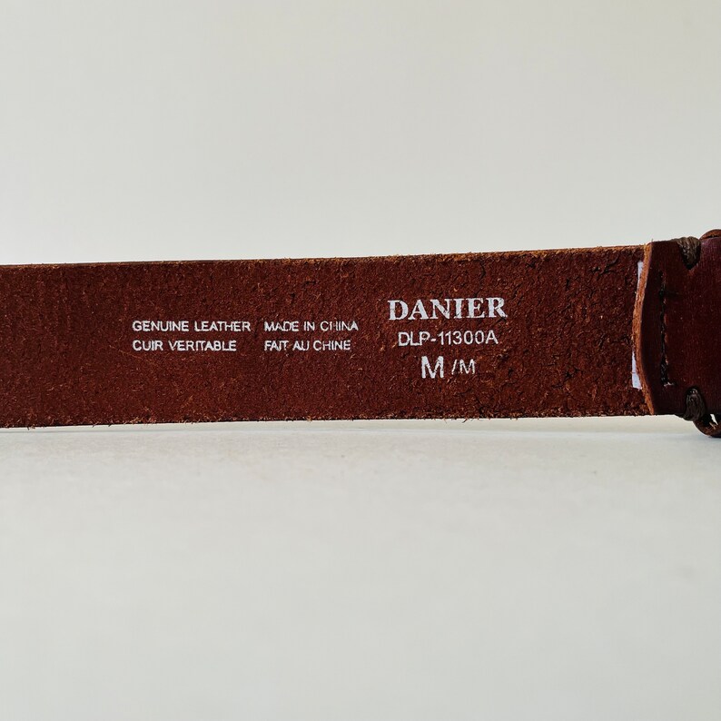 May include: Brown leather belt with white text that reads "GENUINE LEATHER CUIR VERITABLE MADE IN CHINA FAIT AU CHINE DANIER DLP-11300A M/M"