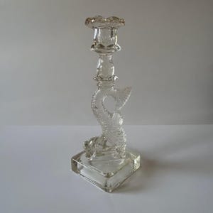 May include: A clear glass candlestick featuring a fish design. The candlestick has a square base and a tall, slender stem. The fish is curled around the stem, with its tail pointing upwards. The candlestick is ideal for adding a touch of elegance to any home.