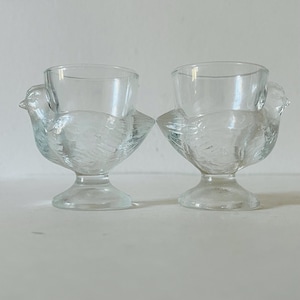 May include: Two clear glass egg cups, each shaped like a bird. The egg cups have a round bowl for the egg and a bird-shaped base with a textured design. The cups are on a white surface.