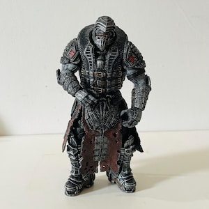 Puede incluir: Figura de acción detallada de un guerrero con armadura gris oscuro con detalles rojos. La figura tiene un casco con una cara amenazante, un gran cuello y una falda deshilachada de color marrón rojizo. La figura está de pie.