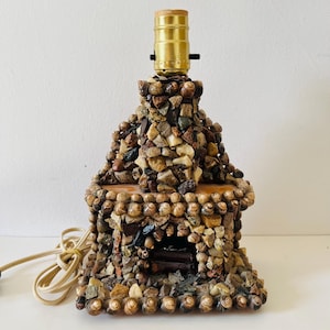 Könnte beinhalten: Eine einzigartige Tischlampe im Kamin-Design, gefertigt aus verschiedenen Steinen und Muscheln. Sie hat eine Messing-Lampenfassung und ein beiges Stromkabel. Der Lampensockel ist rechteckig, und der Kamin ist konisch.