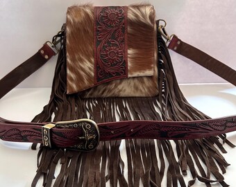 May include: A brown leather crossbody bag with a fringe trim and a cowhide flap. The bag has a brown leather strap with a metal buckle and a floral design.