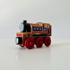 May include: A wooden toy train engine featuring a black front with a face, red wheels, and a tan body. The side displays the text NWR 37, a blue ribbon icon, and blue bunting decorations. The design includes red and black accents on the wooden frame.