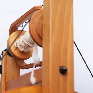 May include: A wooden spinning wheel with a light-coloured wood finish. The wheel has a spool of white yarn with red accents. Black cords and a black metal component are also visible.