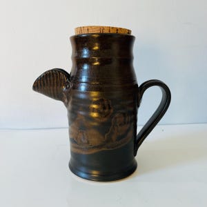 Może przedstawiać: Ręcznie wykonany ceramiczny dzbanek z ciemnobrązową glazurą i korkiem. Dzbanek posiada dzióbek, uchwyt i teksturowany wzór. Ta ceramika to unikalny element wyposażenia kuchni.