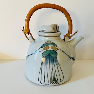 Op de afbeelding: Witte keramische theepot met een bruine houten handgreep en deksel. De theepot is versierd met een abstract blauw, groen en goudkleurig ontwerp. De handgreep is bevestigd met leren riemen.