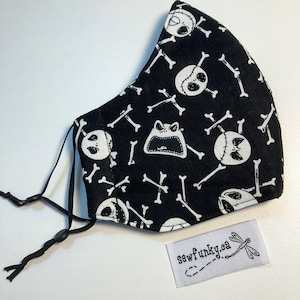 Könnte beinhalten: Schwarze Gesichtsmaske mit einem sich wiederholenden Muster aus weißen Totenköpfen, gekreuzten Knochen und einem schreienden froschartigen Charakter. Die Maske hat verstellbare schwarze elastische Ohrschlaufen und ein kleines weißes Etikett mit dem Text "sewfunky.ca".