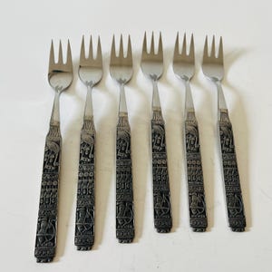 May include: Set of six stainless steel forks with ornate, dark gray handles. The handles feature a detailed, repeating pattern of figures and symbols. The tines are polished and reflect the light. The forks are arranged on a white surface.
