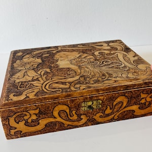 May include: A rectangular wooden box with a detailed carved design. The lid features a profile of a woman with flowing hair and floral elements. The box has a brass clasp and is stained in shades of brown.