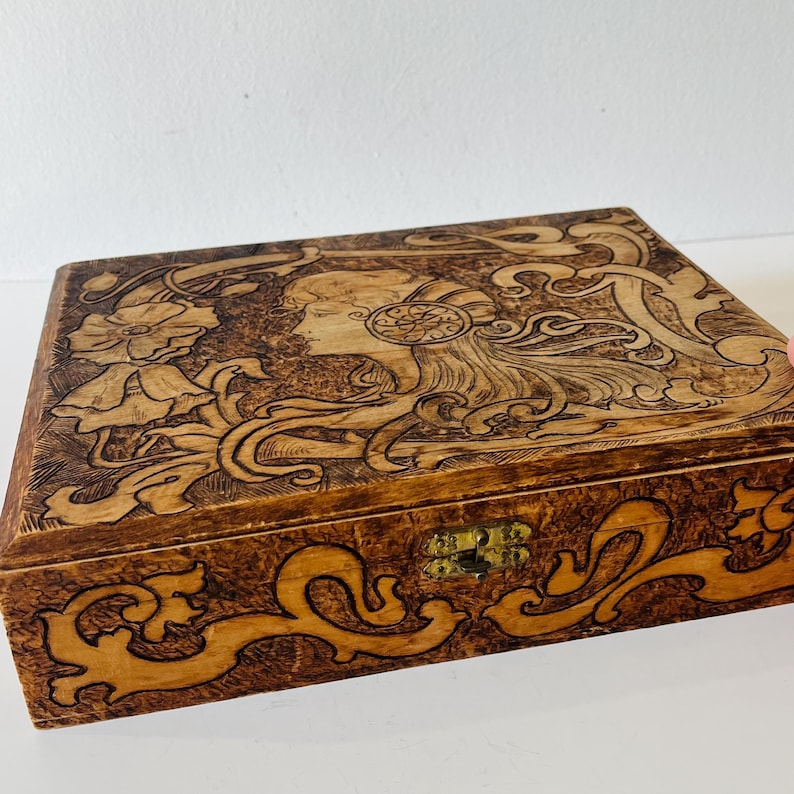 May include: A rectangular wooden box with a detailed carved design. The lid features a profile of a woman with flowing hair and floral elements. The box has a brass clasp and is stained in shades of brown.