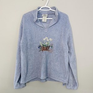 May include: A light blue fleece pullover with a half-zip collar. The front features an embroidered design of a wooden wheelbarrow filled with flowers. The pullover has long sleeves and a drawstring at the hem.