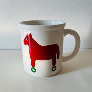 May include: White ceramic mug featuring a red Dala horse design. The horse has a blue eye and green and yellow wheels. The mug has a curved handle and is suitable for hot or cold beverages.