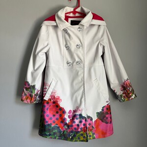 May include: White double-breasted coat with a pink lining and a floral print on the bottom hem. The coat has four buttons and a label that says "Couture".
