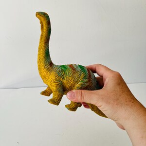 Puede incluir: Un juguete de dinosaurio Braquiosaurio amarillo y verde con cuello largo y patas peque&ntilde;as. El juguete tiene una superficie texturizada y est&aacute; sostenido por una mano.