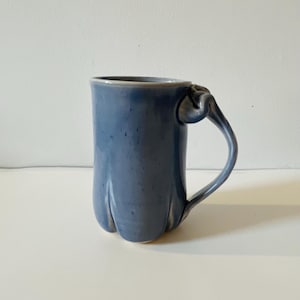 May include: A blue ceramic mug with a unique, sculpted handle. The mug has a textured, slightly speckled glaze.