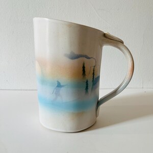 May include: A white ceramic mug featuring a soft, airbrushed landscape design in shades of blue, orange, and tan. The illustration depicts a silhouette of a person skiing through a snowy forest with pine trees and a faint, wispy cloud above.
