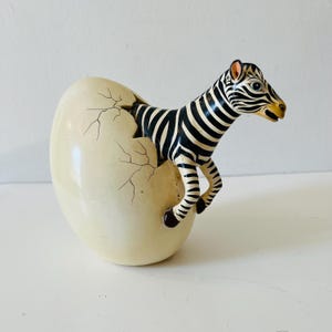 May include: A black and white zebra figurine emerging from a cracked white egg. The zebra has a black mane and a black and white striped body.
