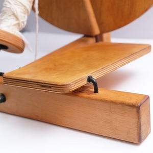 May include: A wooden spinning wheel with a light wood finish. The wheel has a large circular wheel, a foot pedal, and a wooden base. A spool of white yarn is attached to the wheel. The spinning wheel is designed for textile crafts, often used for making yarn.