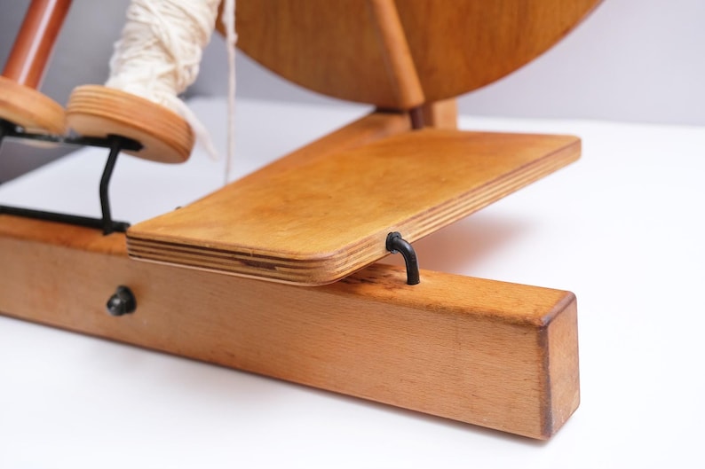 May include: A wooden spinning wheel with a light wood finish. The wheel has a large circular wheel, a foot pedal, and a wooden base. A spool of white yarn is attached to the wheel. The spinning wheel is designed for textile crafts, often used for making yarn.