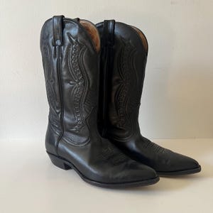 May include: A pair of black leather cowboy boots featuring a pointed toe, a stacked heel, and intricate tonal stitching patterns along the tall shaft. The boots have pull tabs at the top and a smooth finish, showcasing a classic western silhouette.