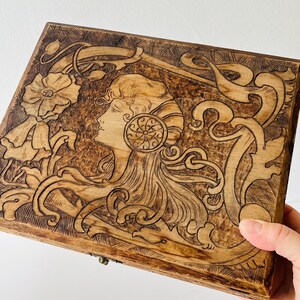 May include: Wooden box with Art Nouveau style carving. The design features a woman's profile, flowers, and flowing lines. The box is a warm brown color and has a hinged lid.