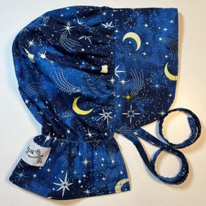 May include: A blue fabric bonnet with a white star and crescent moon pattern. The bonnet has a gathered ruffle at the top and ties at the back.