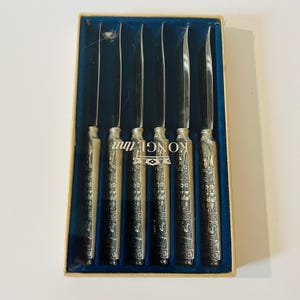 May include: Set of six vintage silver-toned steak knives in a blue-lined box. The handles are intricately detailed with Egyptian-style hieroglyphic designs. The box has the word "KONGE" printed on it.