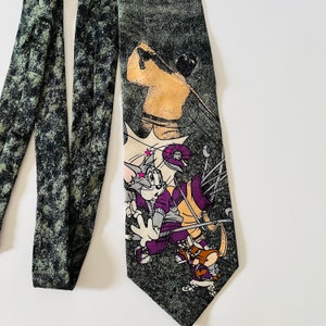May include: A green and black tie with a cartoon image of Tom and Jerry playing golf. Tom is swinging a golf club and Jerry is holding a golf bag. The tie has a distressed, faded look.