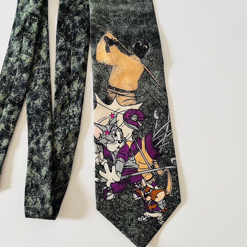 May include: A green and black tie with a cartoon image of Tom and Jerry playing golf. Tom is swinging a golf club and Jerry is holding a golf bag. The tie has a distressed, faded look.